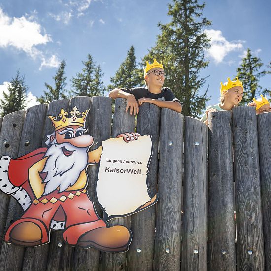 Drei Kinder mit gelben Kronen blicken über den hölzernen Zaun am Eingang zur KaiserWelt, daneben ein buntes Schild mit gezeichnetem König und der Aufschrift Eingang | entrance KaiserWelt