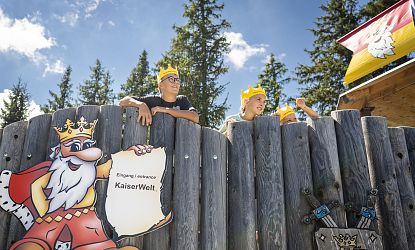 Drei Kinder mit gelben Kronen blicken über den hölzernen Zaun am Eingang zur KaiserWelt, daneben ein buntes Schild mit gezeichnetem König und der Aufschrift Eingang | entrance KaiserWelt