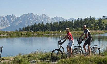Ein Paar fährt mit E-Bikes auf einem Weg entlang eines ruhigen Sees, im Hintergrund erheben sich die schroffen Gipfel des Wilden Kaisers