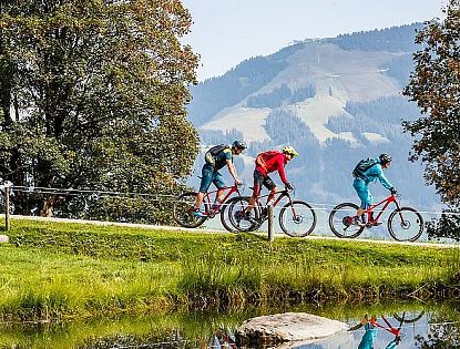 Drei Mountainbiker fahren auf einem Schotterweg entlang eines kleinen Sees in einer grünen Berglandschaft mit Bäumen und Blick auf bewaldete Hügel im Hintergrund