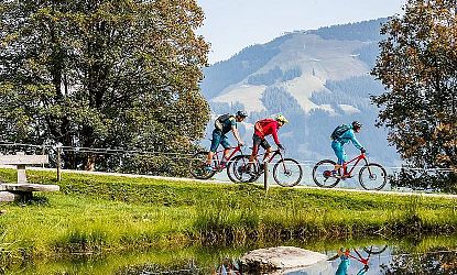 Drei Mountainbiker fahren auf einem Schotterweg entlang eines kleinen Sees in einer grünen Berglandschaft mit Bäumen und Blick auf bewaldete Hügel im Hintergrund
