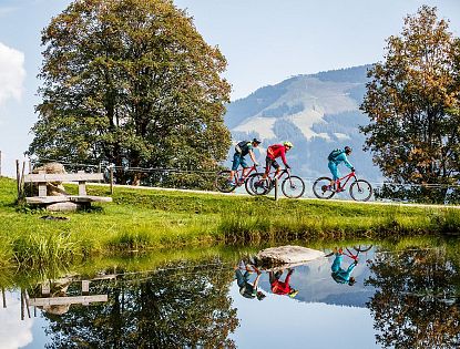 Drei Mountainbiker fahren auf einem Schotterweg entlang eines kleinen Sees in einer grünen Berglandschaft mit Bäumen und Blick auf bewaldete Hügel im Hintergrund