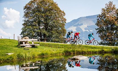 Drei Mountainbiker fahren auf einem Schotterweg entlang eines kleinen Sees in einer grünen Berglandschaft mit Bäumen und Blick auf bewaldete Hügel im Hintergrund