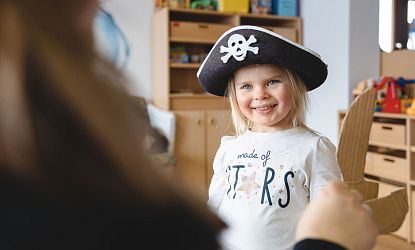 Lächelndes Mädchen mit Piratenhut und Kartonschwert steht in einem Raum mit Holzregalen und Spielzeug.