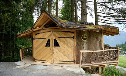 Märchenhafte Holzhütte, die Kunstwerker Hütte im Hexenwald Hexenwasser mit schrägem Dach, Rundfenster und Ast-Geländer, eingebettet zwischen Bäumen am Waldrand mit Blick auf die Berge.