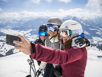 Drei Skifahrerinnen am Ladies Day mit Helmen und Skibrillen machen lachend ein Selfie auf einer verschneiten Piste mit weitem Blick über die winterliche Berglandschaft