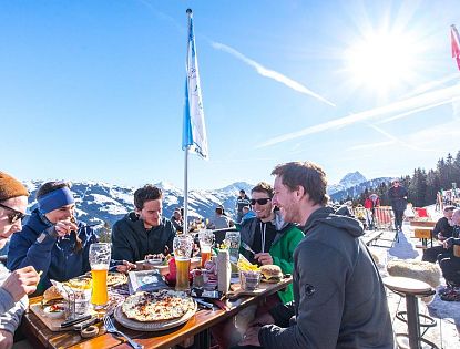 Gruppe junger Menschen genießt Pizza und Getränke an einem Holztisch im Freien auf einer verschneiten Sonnenterrasse mit Blick auf schneebedeckte Berge am Mens Day in der SkiWelt