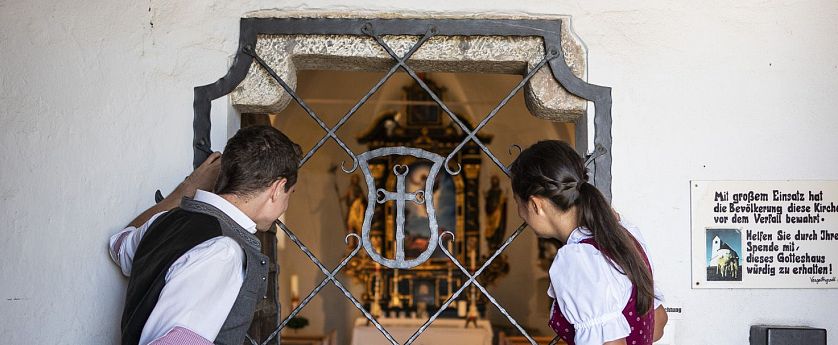 Zwei traditonell gekleidete Personen schauen durch die Eingangstüre der Salvenkirche auf der Hohen Salve.