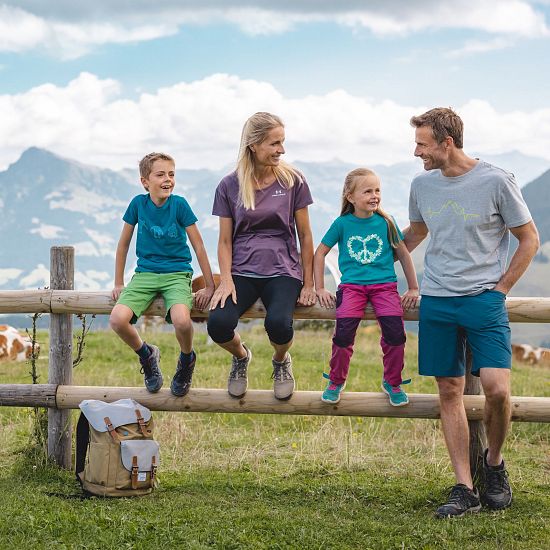 Familie beim Wandern macht eine Pause sitz auf einem Holzbalken eines Zaunes, Rucksatz steht am Boden und eine Kuh sowie Alpenlanschaft im Hintergrund zu sehen