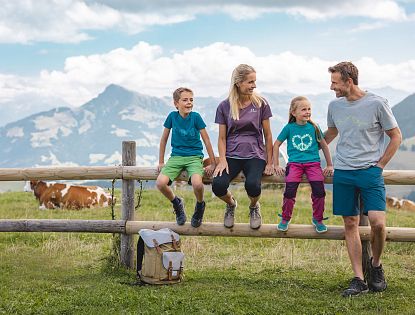 Familie beim Wandern macht eine Pause sitz auf einem Holzbalken eines Zaunes, Rucksatz steht am Boden und eine Kuh sowie Alpenlanschaft im Hintergrund zu sehen